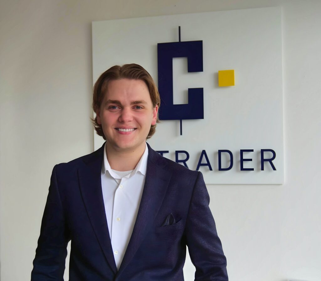 Philip Weihrauch mit schulterlangem Haar, die einen dunklen Blazer und ein weißes Hemd trägt, steht vor einem Schild mit der Aufschrift „TRADER“.