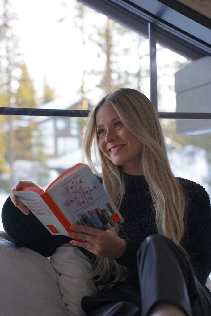 Eine Person mit langen blonden Haaren sitzt lächelt auf einem Sofa und liest ein Buch mit dem Titel „Felix mit dastübi Geld“ über Liebe und Geld.