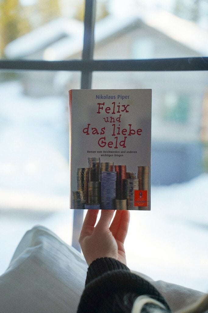 Eine Person hält ein Buch mit dem Titel „Felix und das liebe Geld“ vor einer verschneiten Fensterszene. Auf dem Buchcover ist ein Stapel Münzen zu sehen, der die Essenz von LIEBE und GELD verkörpert.