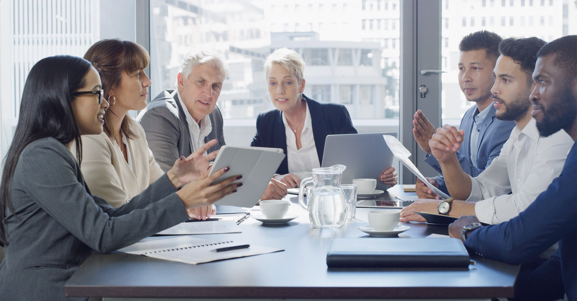 A group of business people are sitting around a conference table and discussing. They are using tablets and laptops. There are cups and a jug of water on the table.