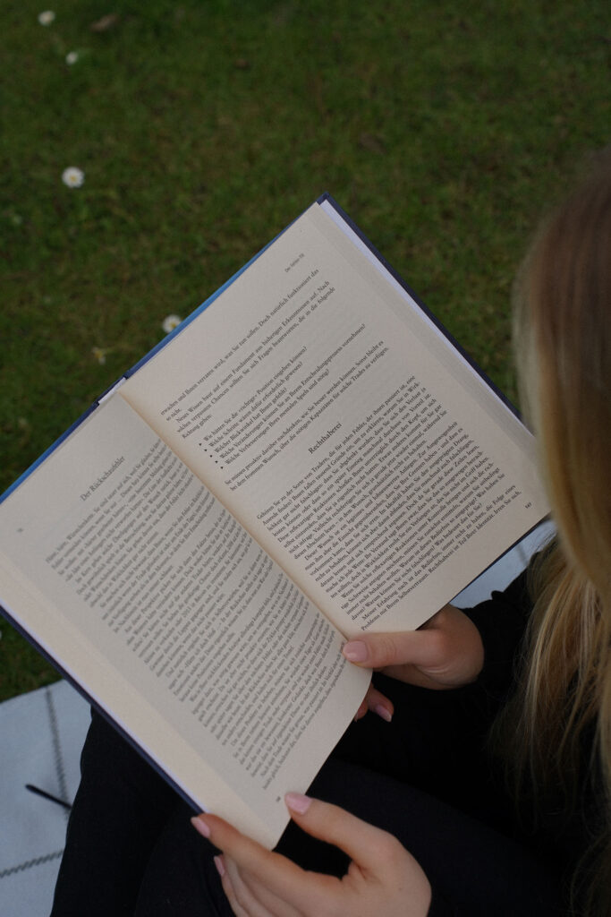 Eine Person hält im Freien auf einer Grasfläche ein offenes Buch in der Hand und liest es. Die mit Text gefüllten Seiten scheinen einem automatisch gespeicherten Entwurf zu entsprechen.