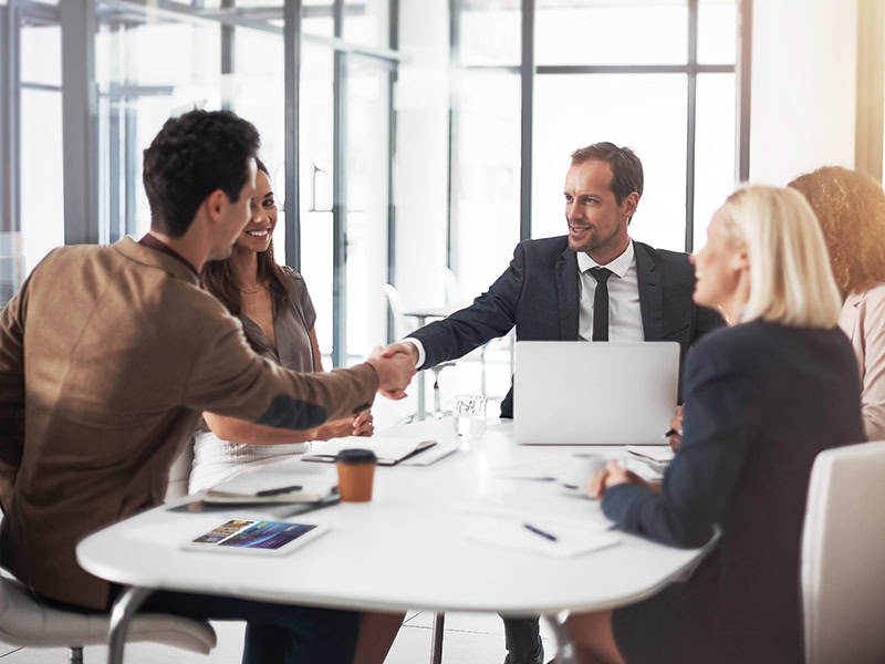 People in business attire sit around a conference table at an office meeting, with two people shaking hands and others looking on. There is also a laptop and a tablet on the table.