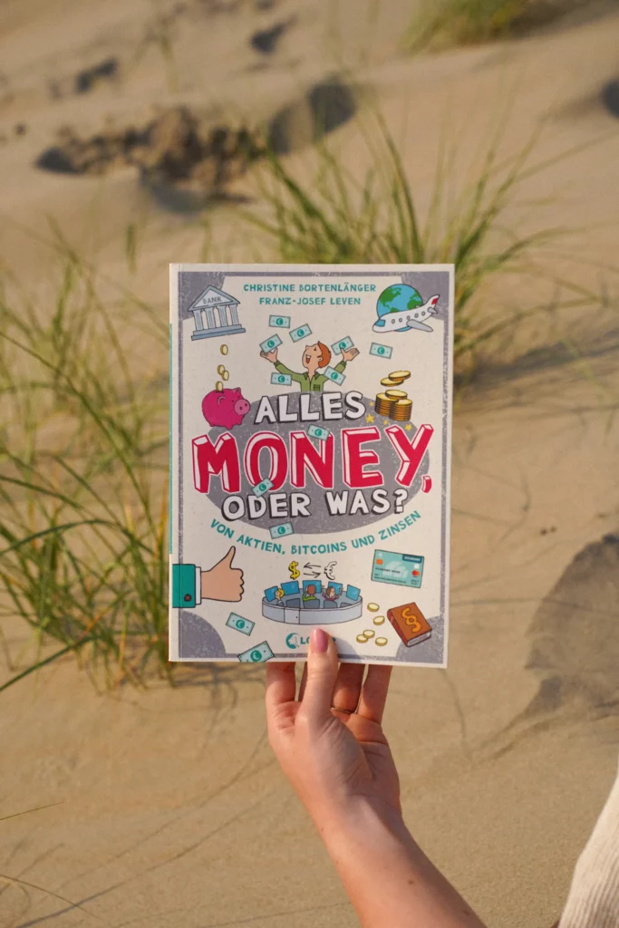 Eine Person hält ein Buch mit dem Titel „Alles Geld Oder Was? Von Aktien, Bitcoins und Zinsen“ vor einem sandigen Hintergrund mit etwas Gras.