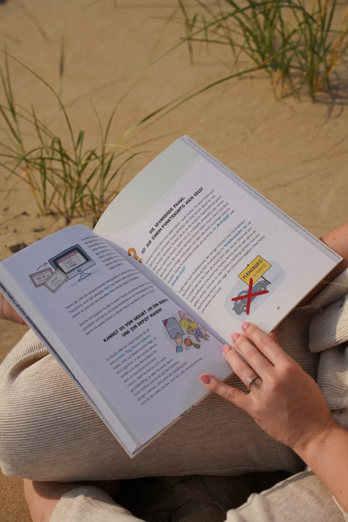 Eine Person sitzt auf Sand und Gras und hält ein offenes Buch in der Hand. Das Buch enthält Abbildungen, darunter einen Computerbildschirm und ein Schild mit durchgestrichenem „Nein“.