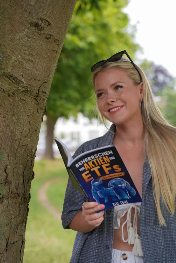 Eine Person mit langen blonden Haaren steht im Freien, lächelt und hält ein aufgeschlagenes Buch mit dem Titel „Beneerschen zu Aktien und ETFs“. Sie ist leger gekleidet und trägt eine Sonnenbrille auf dem Kopf.