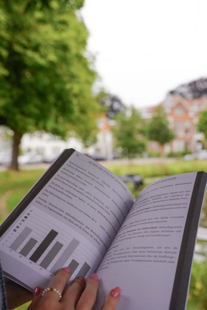 Eine Person hält im Freien ein offenes Buch mit sichtbaren Grafiken und Textseiten. Bäume und Gebäude sind im Hintergrund verschwommen.