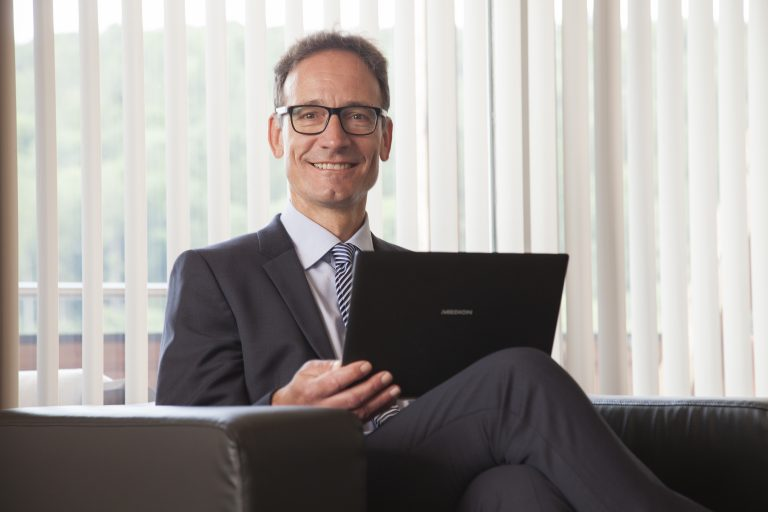 Christian Schwarzkopf in Anzug und Krawatte sitzt lächelnd auf einem Sofa und hält einen aufgeklappten Laptop in der Hand. Vertikale Jalousien im Hintergrund lassen Tageslicht in den Raum.