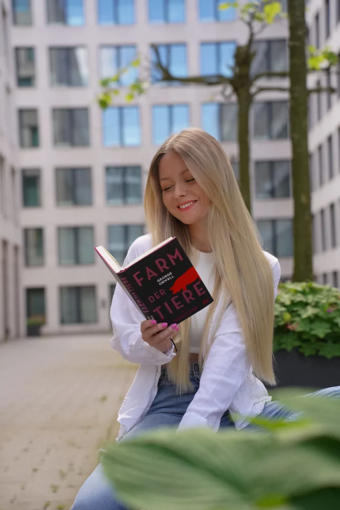 Eine Frau mit langen blonden Haaren sitzt draußen vor einem modernen Gebäude mit zahlreichen Fenstern und liest ein Buch.