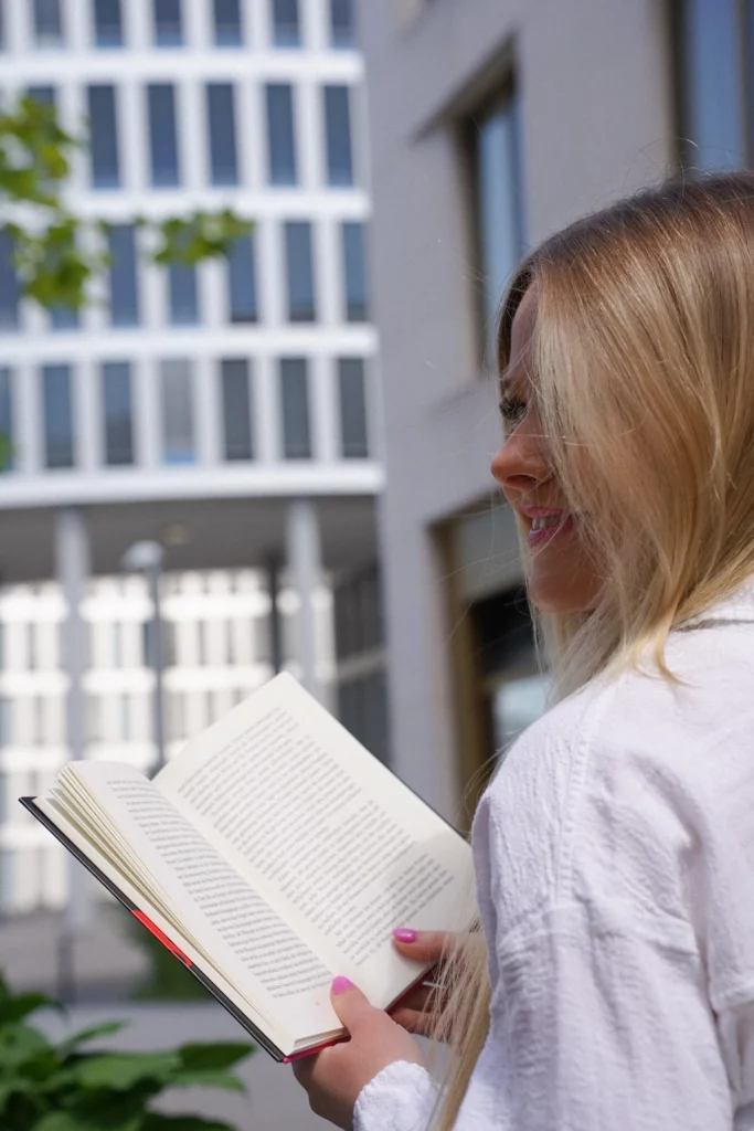 Eine Frau mit langen blonden Haaren und einem weißen Oberteil liest ein offenes Buch, während sie draußen in der Nähe von Gebäuden steht.