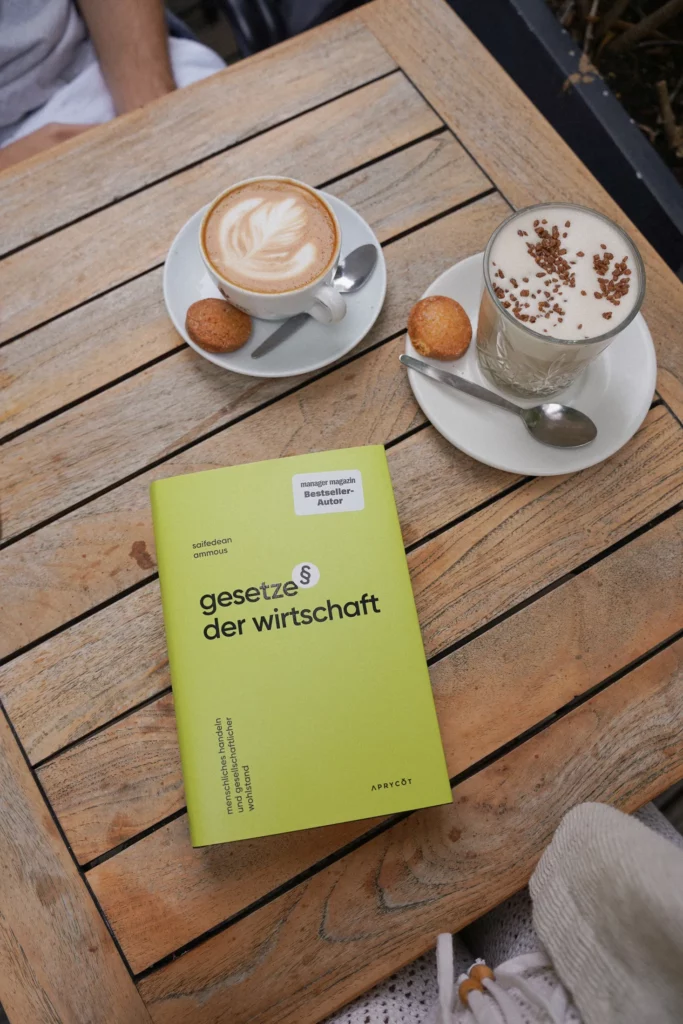 Ein Holztisch mit zwei Tassen Kaffee, eine Cappuccino, eine Latte mit Schlagsahne, jeweils mit einem kleinen Keks. Daneben liegt ein grünes Buch mit dem Titel „Gesetze der Wirtschaft“.