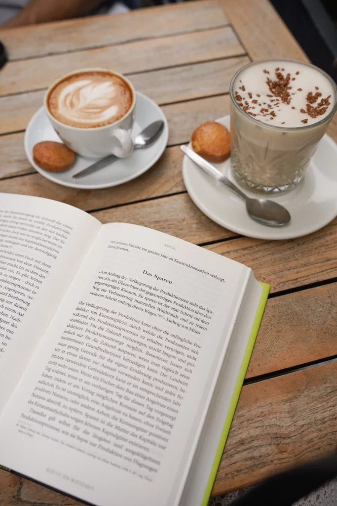 Ein aufgeschlagenes Buch auf einem Tisch neben zwei Tassen Kaffee; eine hat schaumige Latte Art, die andere ist mit Schokostreuseln garniert. Zu jeder Tasse gibt es einen kleinen runden Keks.