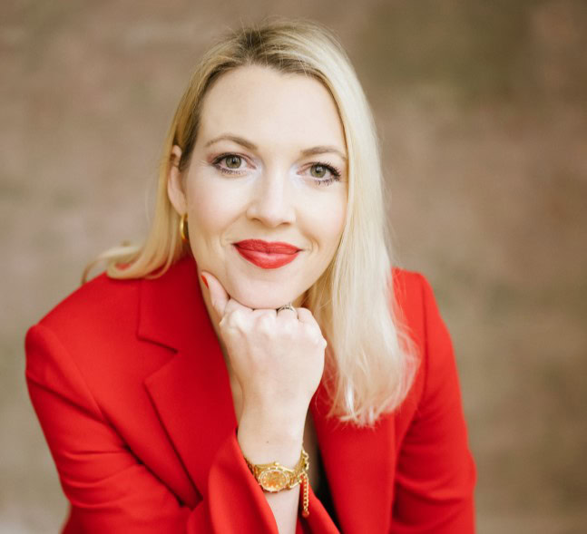 Isabel Poier-Kisling mit blondem Haar, einem roten Blazer und einer goldenen Uhr trägt, stützt ihr Kinn auf ihre Hand und blickt vor einem schlichten Hintergrund direkt in die Kamera.
