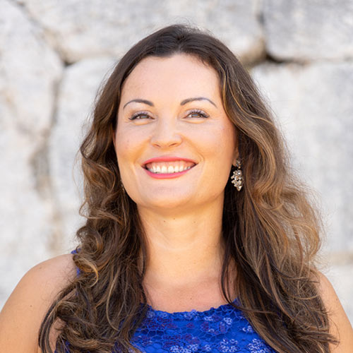 Jana Misar with long, wavy brown hair and a blue lace top smiles against a stone background.