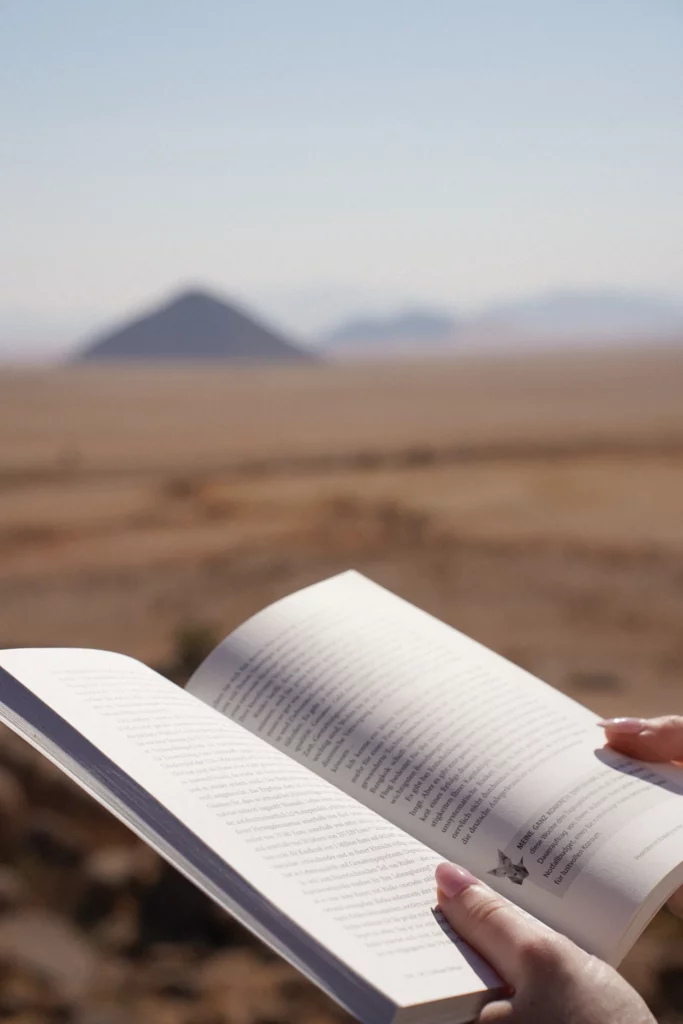 Eine Person liest im Freien in einer Wüstenlandschaft ein Buch. Im Hintergrund ist in der Ferne ein pyramidenförmiger Hügel zu sehen.