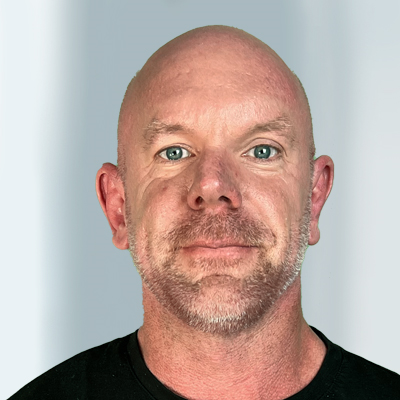 Tradingcoach Martin Goersch mit kurzem Bart und Schnurrbart, der ein schwarzes Hemd trägt und mit neutralem Gesichtsausdruck direkt in die Kamera blickt. Der Hintergrund ist grau und wirkt weich.