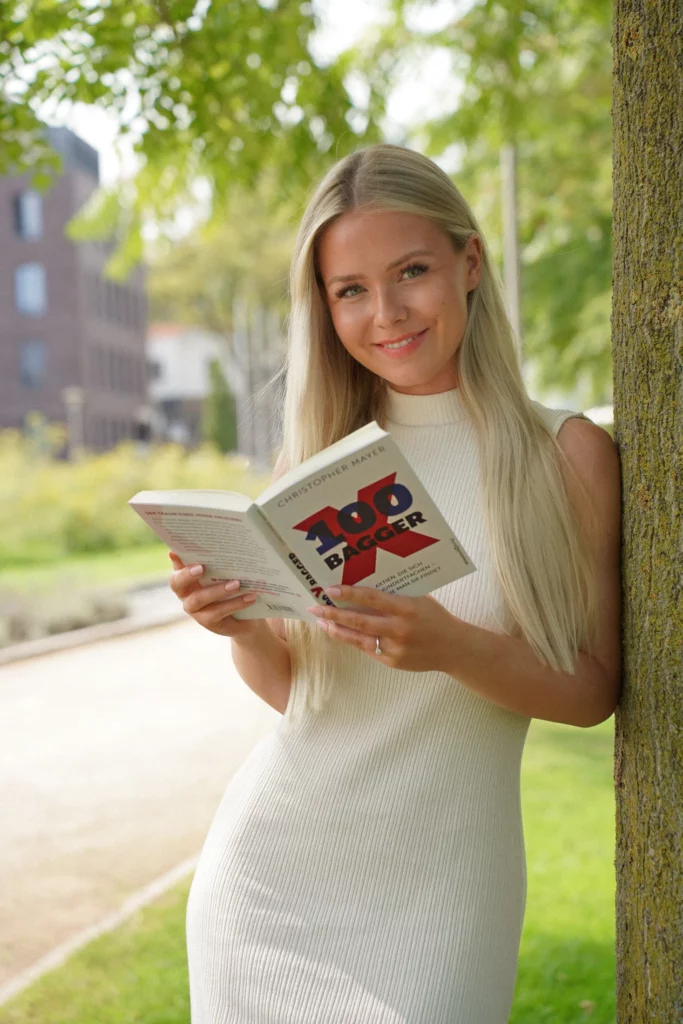 Eine Frau mit langen blonden Haaren und einem weißen ärmellosen Kleid steht im Freien neben einem Baum, hält ein Buch mit dem Titel „100 Baggers“ in der Hand und liest es. Ihre gelassene Haltung lenkt automatisch die Aufmerksamkeit von der Umgebung auf sich.