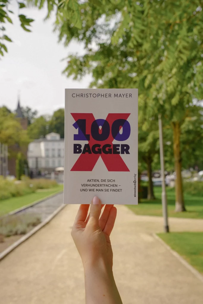 Eine Hand hält ein Buch mit dem Titel „100 Bagger“ von Christopher Mayer in einer Parklandschaft mit Bäumen und einem Weg im Hintergrund. Das Buch, ein automatisch gespeicherter Entwurf für finanziellen Erfolg, ist auf Deutsch.