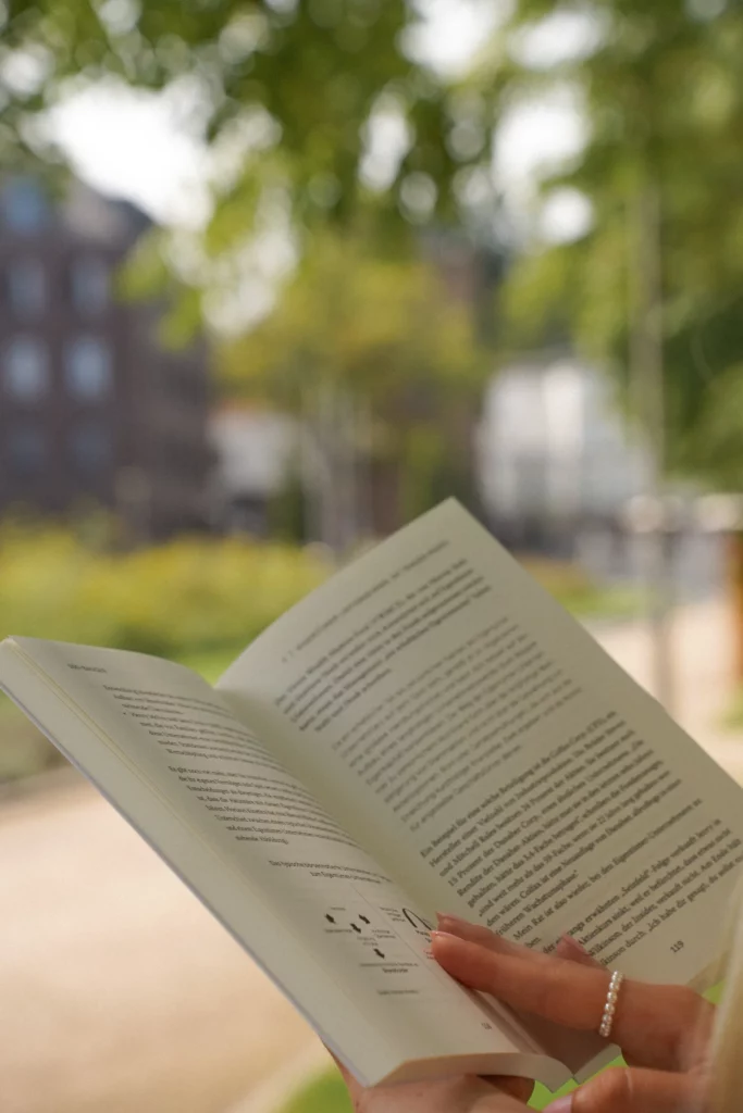 Eine Person liest im Freien ein Buch mit verschwommenem Grün und Gebäuden im Hintergrund, das fast wie ein automatisch gespeicherter Entwurf aussieht.
