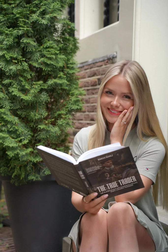 Frau mit langen blonden Haaren sitzt im Freien, lächelt und hält ein offenes Buch mit dem Titel „The True Trader“. Im Hintergrund steht eine Topfpflanze, die die Szene wie einen automatisch gespeicherten Entwurf vervollständigt.