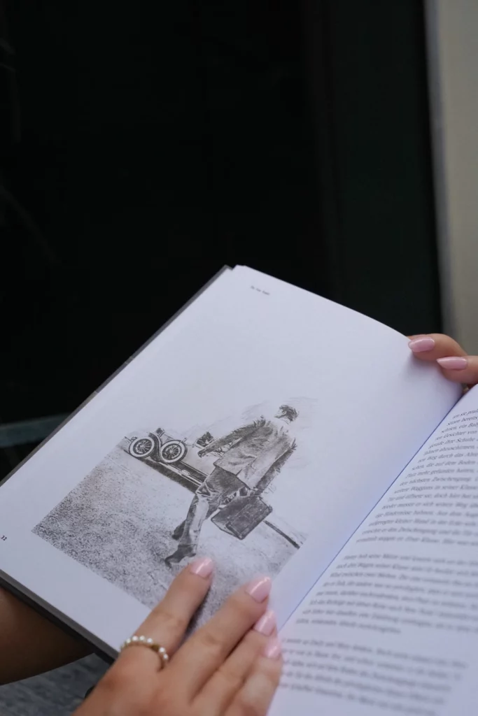 Eine Person mit manikürten Nägeln hält ein offenes Buch in der Hand. Darin ist die Schwarzweißzeichnung eines Mannes zu sehen, der mit einem Koffer in der Hand von einem Oldtimer weggeht, als wäre sie in einem automatisch gespeicherten Entwurf festgehalten.