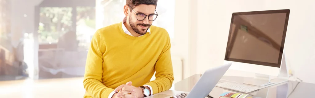 Ein Mann mit Brille und gelbem Pullover sitzt an einem Schreibtisch und schaut auf einen Laptop. Auf dem Schreibtisch in einem hellen, sonnendurchfluteten Raum liegen ein Desktop-Computer und bunte Papiere.
