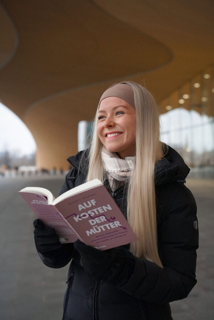 Person mit langen blonden Haaren lächelt, während sie vor einem modernen Gebäude ein offenes Buch mit dem Titel „Auf Kosten der Mutter“ hält.