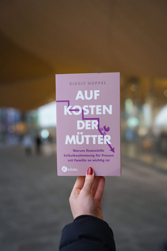 Hand hält ein Buch mit dem Titel „Auf Kosten der Mütter“ von Birgit Happel, mit verschwommenem Stadthintergrund.