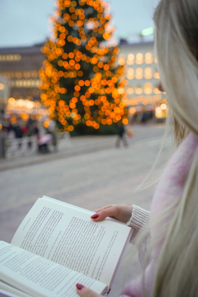 Person liest ein Buch vor einem großen, mit Lichtern geschmückten Weihnachtsbaum im Freien.