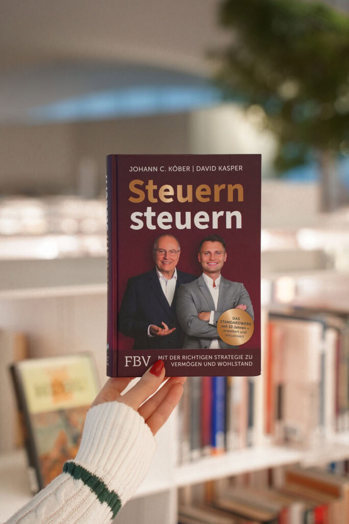 Person hält ein Buch mit dem Titel „Steuern steuern“ von Johann C. Köber und David Kasper in einer Bibliothek.