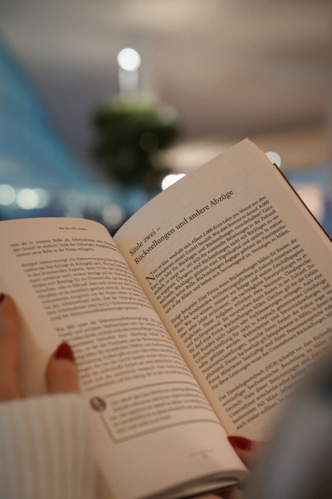 Eine Person mit roten Nägeln hält ein aufgeschlagenes Buch in deutscher Sprache. Im Hintergrund ist eine verschwommene Pflanze zu sehen.