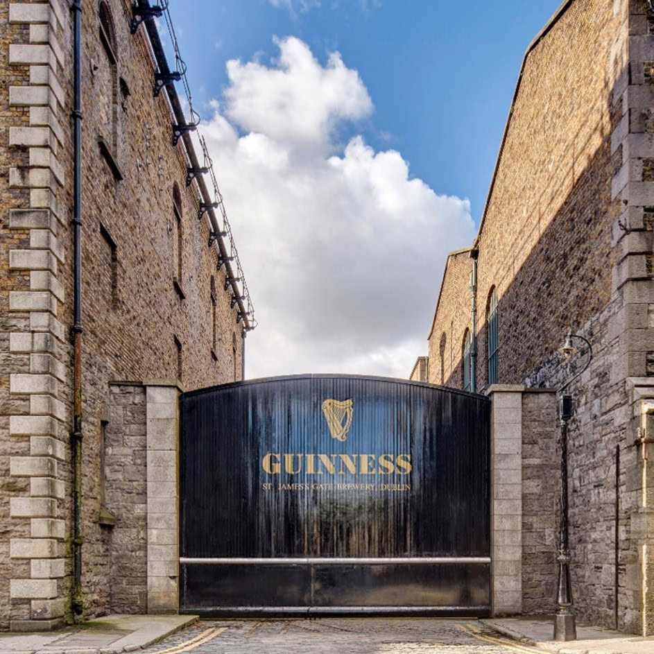 Large black gate with "GUINNESS" and logo in gold lettering, flanked by stone walls under a partly cloudy sky.