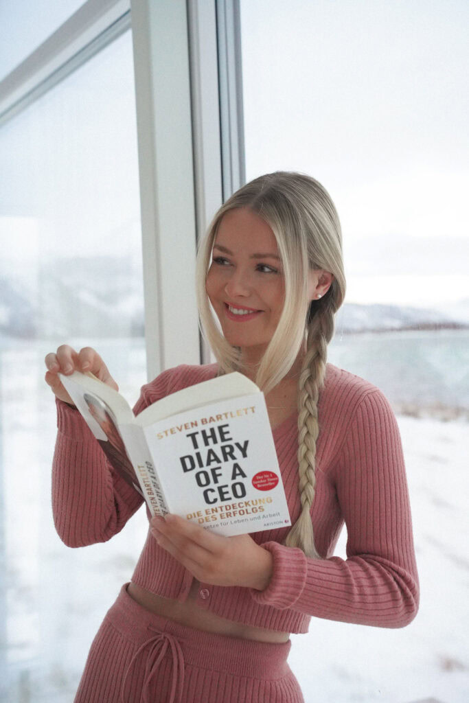 Eine Person mit einem langen blonden Zopf und einem rosa Outfit steht im Innenbereich an einem Fenster und hält ein Buch mit dem Titel „Das Tagebuch eines CEOs“. Draußen ist eine verschneite Landschaft zu sehen.