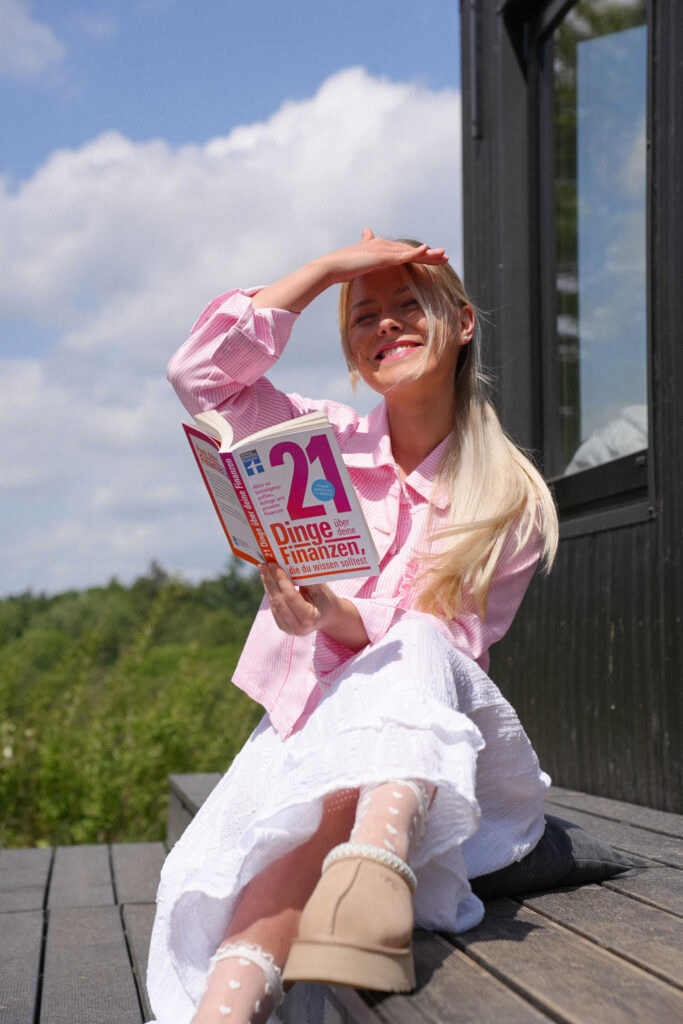 Eine Frau sitzt im Freien auf Holzstufen, hält ein Buch mit dem Titel „21 Dinge, die du über Finanzen wissen musst“ in der Hand und schützt ihre Augen mit der Hand vor der Sonne.
