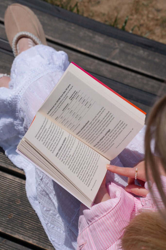 Eine Person in einem rosa Hemd und einem weißen Rock sitzt im Freien auf einer Holzfläche und liest ein offenes Buch, das Text und eine Tabelle mit Daten anzeigt.