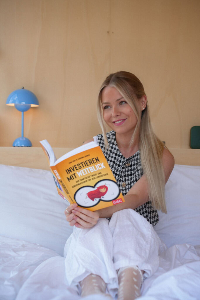 Eine Frau sitzt lächelnd auf einem Bett und liest ein Buch mit dem Titel „Investieren mit Weitblick“. Im Hintergrund ist eine blaue Lampe zu sehen.