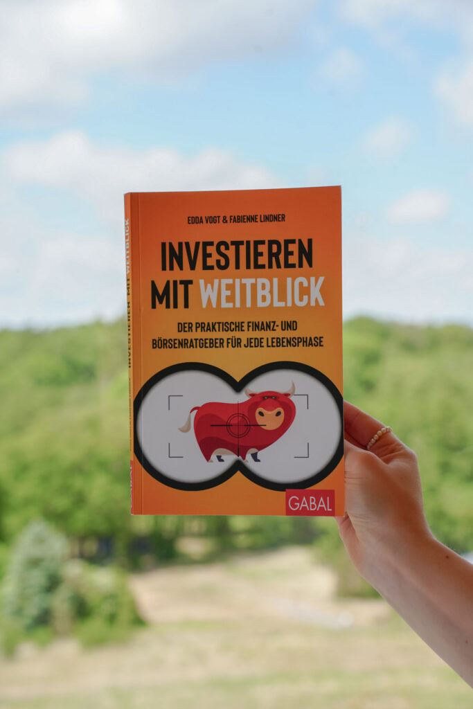 Eine Hand hält das Buch „Investieren mit Weitblick“ von Edda Vogt & Fabienne Lindner vor einem unscharfen Außenhintergrund mit Grün und blauem Himmel.