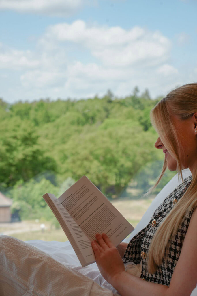 Eine Frau mit langen blonden Haaren liest ein Buch, während sie an einem Fenster mit Blick auf grüne Bäume und einen teilweise bewölkten Himmel sitzt.