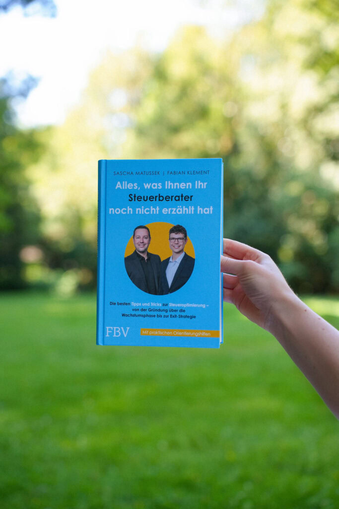 Eine Hand hält ein deutsches Buch mit dem Titel "Alles, was Ihnen Ihr Steuerberater noch nicht erzählt hat" vor einem unscharfen Außenhintergrund mit grünem Gras und Bäumen hoch.