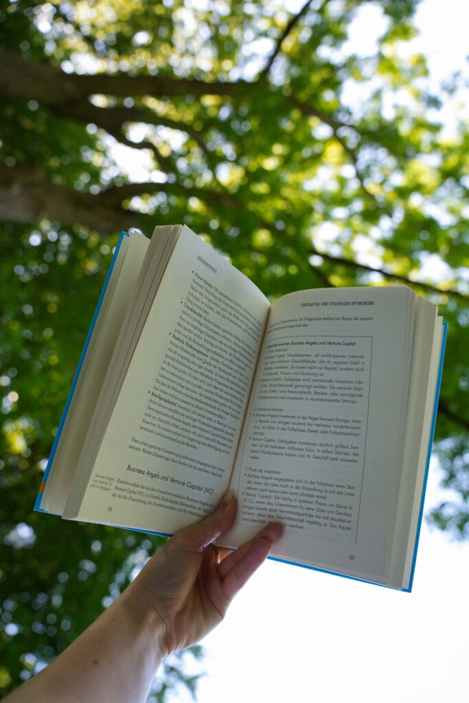 Eine Hand hält ein aufgeschlagenes Buch im Freien mit grünen Baumblättern und Sonnenlicht im Hintergrund.
