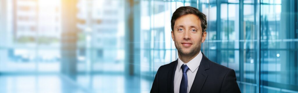 Ein Mann im Geschäftsanzug steht in einem modernen gläsernen Bürogebäude und schaut mit neutralem Blick in die Kamera.