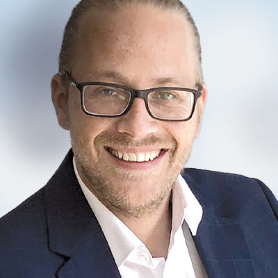 Ein Mann mit Brille und Bart, der einen dunklen Blazer und ein weißes Hemd trägt, lächelt in die Kamera vor einem hellen Hintergrund mit Farbverlauf.