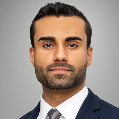 A man with short dark hair, a trimmed beard and a neutral expression, wearing a dark suit, a white shirt and a gray tie, poses in front of a plain gray background.