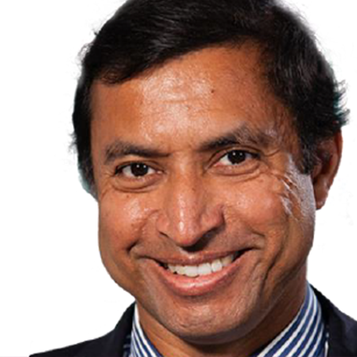 A man with dark hair and brown skin smiles at the camera. He is wearing a dark jacket and a collared shirt with white and blue stripes. The background is white.