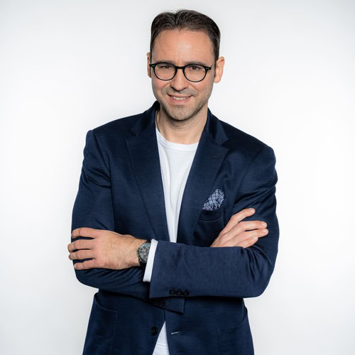 Ein Mann mit Brille und einem marineblauen Blazer über einem weißen Hemd steht mit verschränkten Armen lächelnd vor einem schlichten weißen Hintergrund.