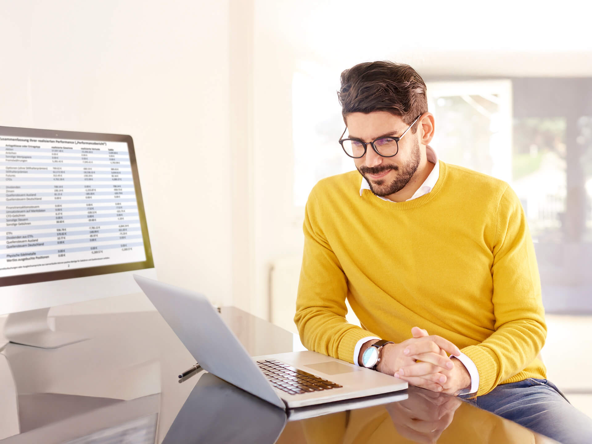 A man in a yellow sweater sits at a desk and looks at a laptop, while a desktop monitor displays a spreadsheet in the background.