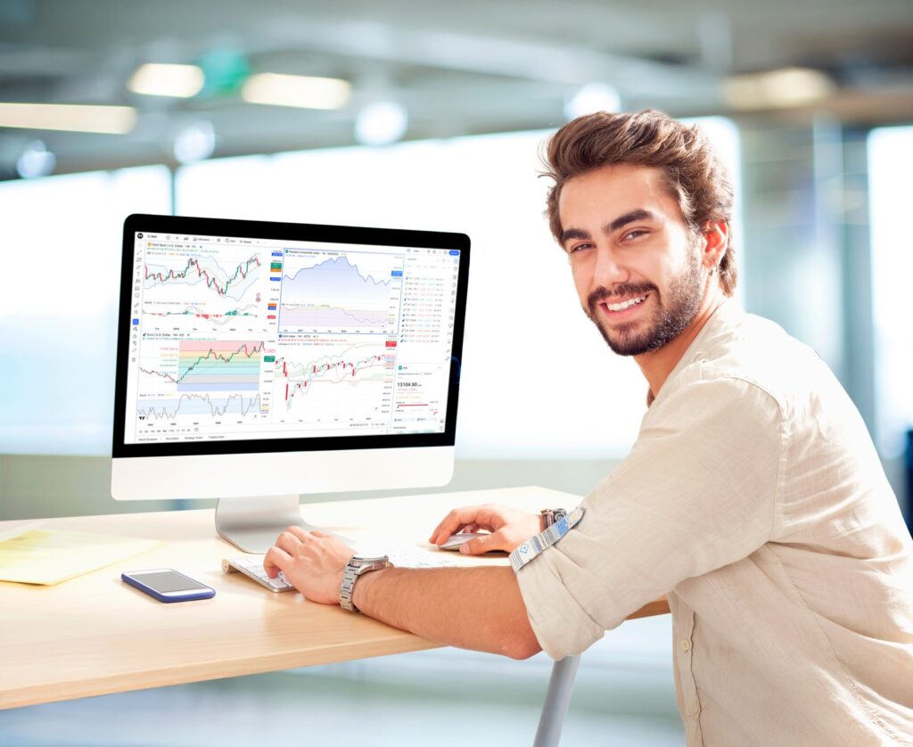 Ein Mann sitzt an einem Schreibtisch in einem Büro und blickt auf einen Computerbildschirm, auf dem mehrere Finanzdiagramme angezeigt werden; neben ihm liegt ein Smartphone.