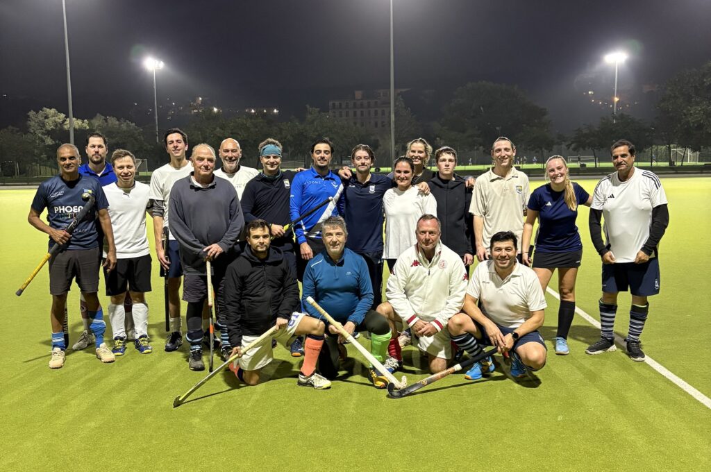 Eine Gruppe von Männern und Frauen posiert nachts auf einem beleuchteten Feldhockeyfeld, einige knien davor, alle halten Hockeyschläger und tragen Sportkleidung - ein inspirierender Teamgeist, ähnlich wie bei langfristig investieren für nachhaltigen Erfolg.