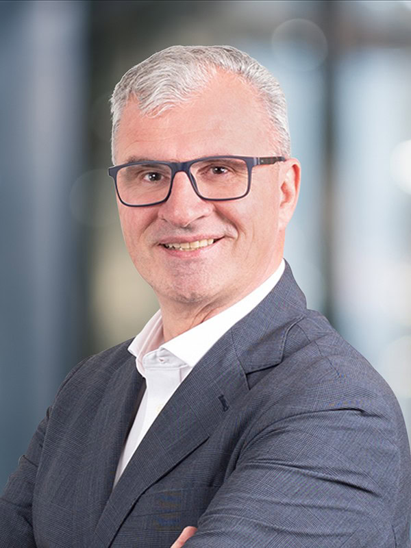 Ein Mann mittleren Alters mit kurzen grauen Haaren, der eine Brille, ein weißes Hemd und einen grauen Blazer trägt, lächelt mit verschränkten Armen vor einem verschwommenen Bürohintergrund.