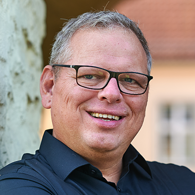 Ein Mann mittleren Alters mit kurzen grauen Haaren und Brille lächelt, trägt ein schwarzes Hemd mit Kragen und steht im Freien an einer strukturierten Wand.