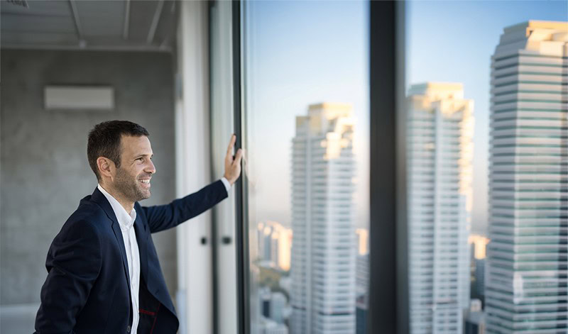 Ein Mann im Anzug steht an einem großen Fenster in einem modernen Büro und blickt auf mehrere hohe Gebäude unter einem klaren Himmel.